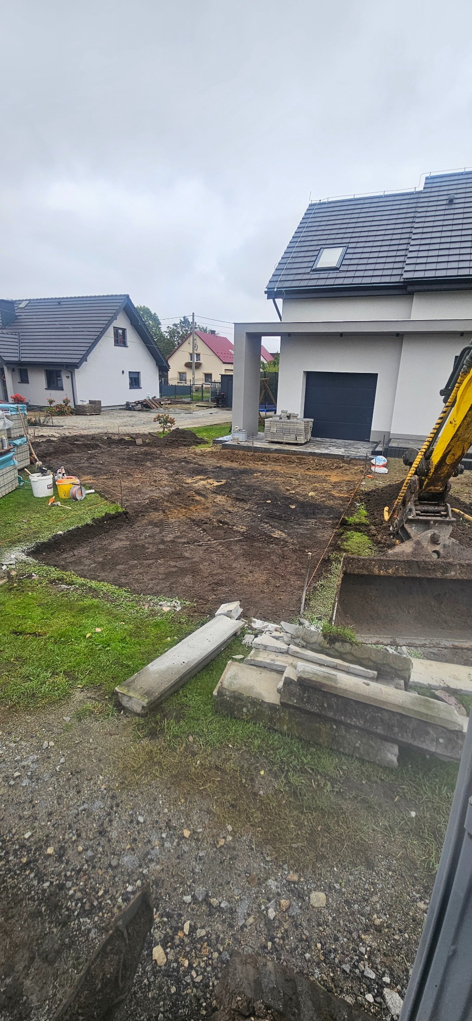 Wykopany teren pod budowę domu jednorodzinnego, widoczny fragment koparki, wyznaczone granice, przygotowane materiały budowlane. Pochmurne niebo.