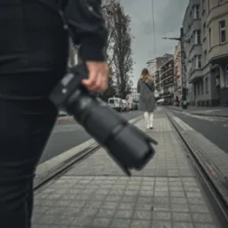 Osoba z aparatem fotograficznym obserwuje kobietę idącą po torach tramwajowych w miejskiej scenerii, sugerując śledzenie lub dyskretną obserwację.