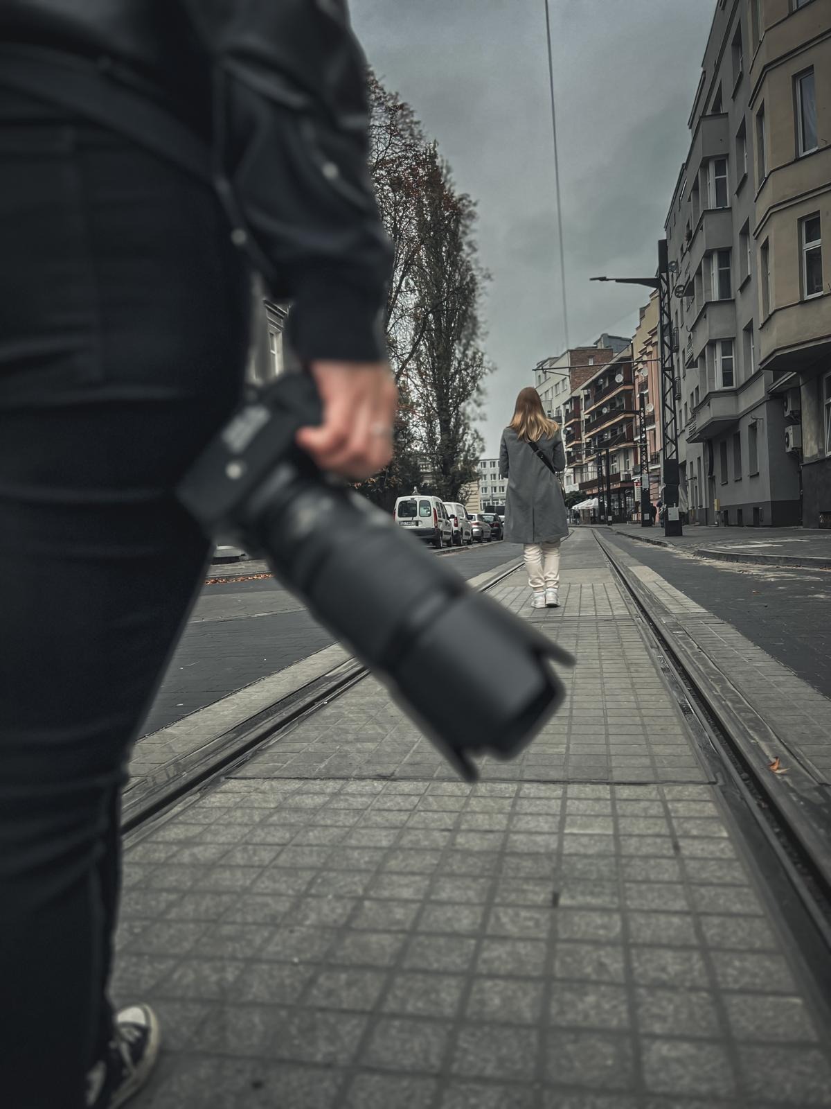 Osoba z aparatem fotograficznym obserwuje kobietę idącą po torach tramwajowych w miejskiej scenerii, sugerując śledzenie lub dyskretną obserwację.