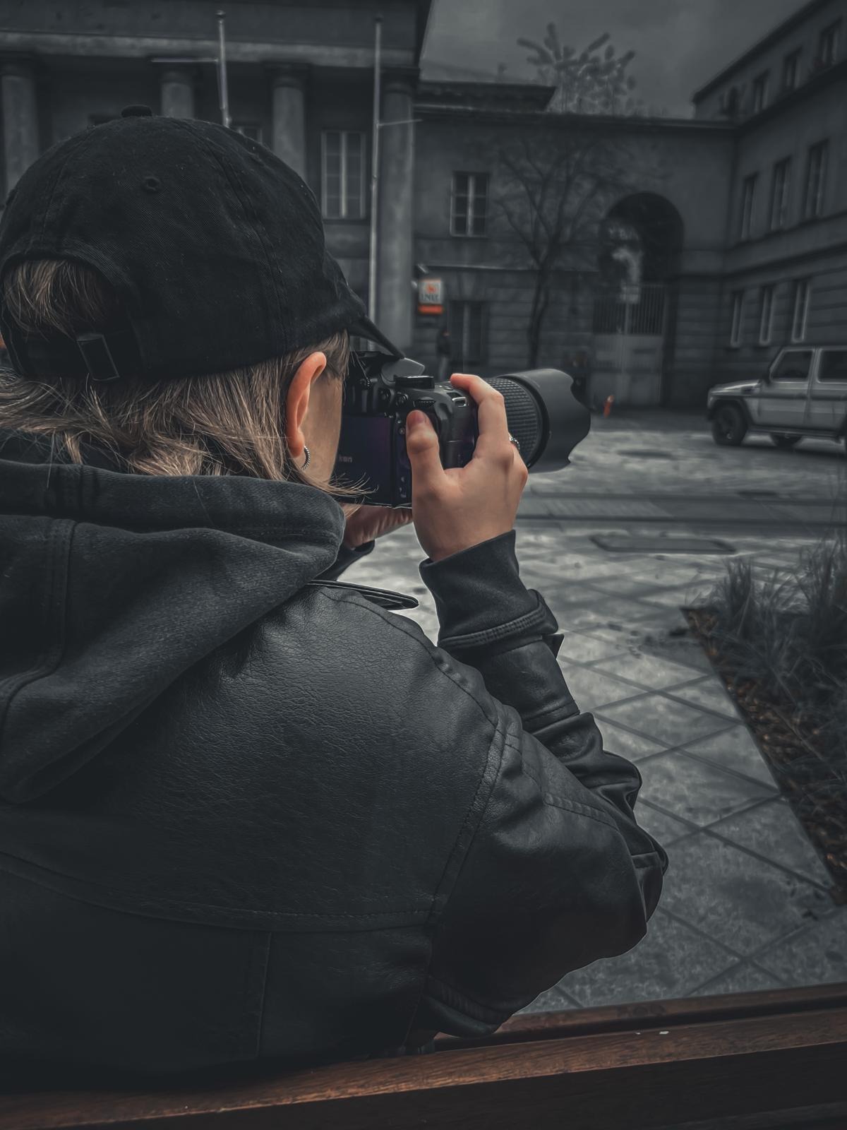 Osoba z aparatem fotograficznym w Sosnowcu, z tyłu budynek z kolumnami i samochód. Kadr zza pleców, ciemna tonacja, widoczny fragment ławki.