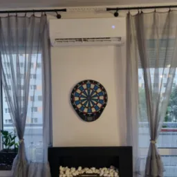 Biała klimatyzacja Rotenso Roni na ścianie między oknami z szarymi zasłonami, poniżej tarcza do darta i dekoracyjny kominek z białymi kamieniami.