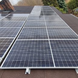 KLIMA SOLUTIONS Łukasz Musiał - Panel fotowoltaiczny na dachu pokrytym papą, z widoczną drabiną i kominem w tle. Widok ukończonej instalacji z bliska, w pochmurny dzień.