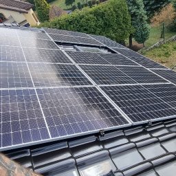 KLIMA SOLUTIONS Łukasz Musiał - Czarny dach z panelami fotowoltaicznymi w słoneczny dzień, widok z góry, na tle domu i zieleni. W tle zaparkowany samochód i opony.