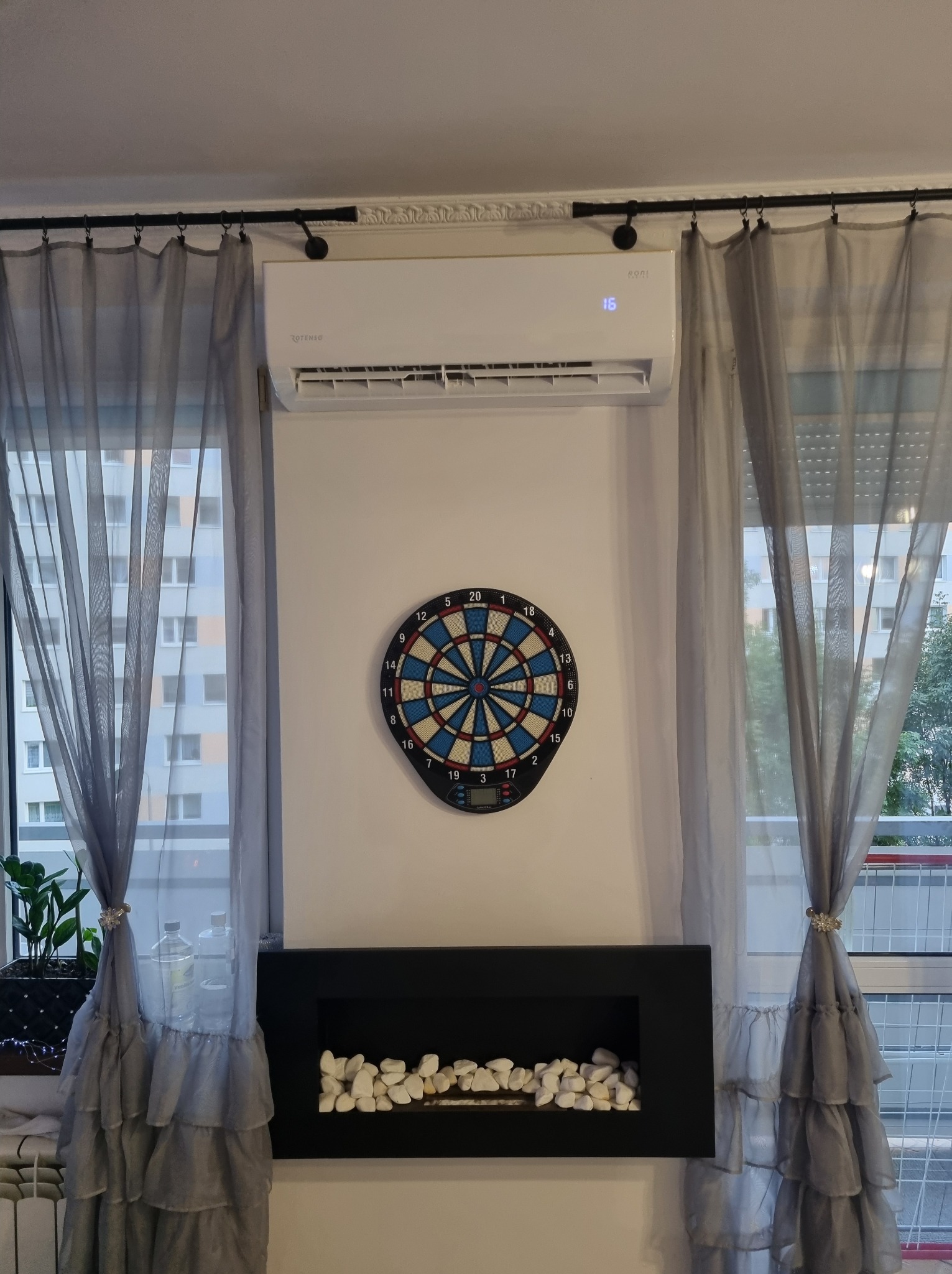 Biała klimatyzacja Rotenso Roni na ścianie między oknami z szarymi zasłonami, poniżej tarcza do darta i dekoracyjny kominek z białymi kamieniami.
