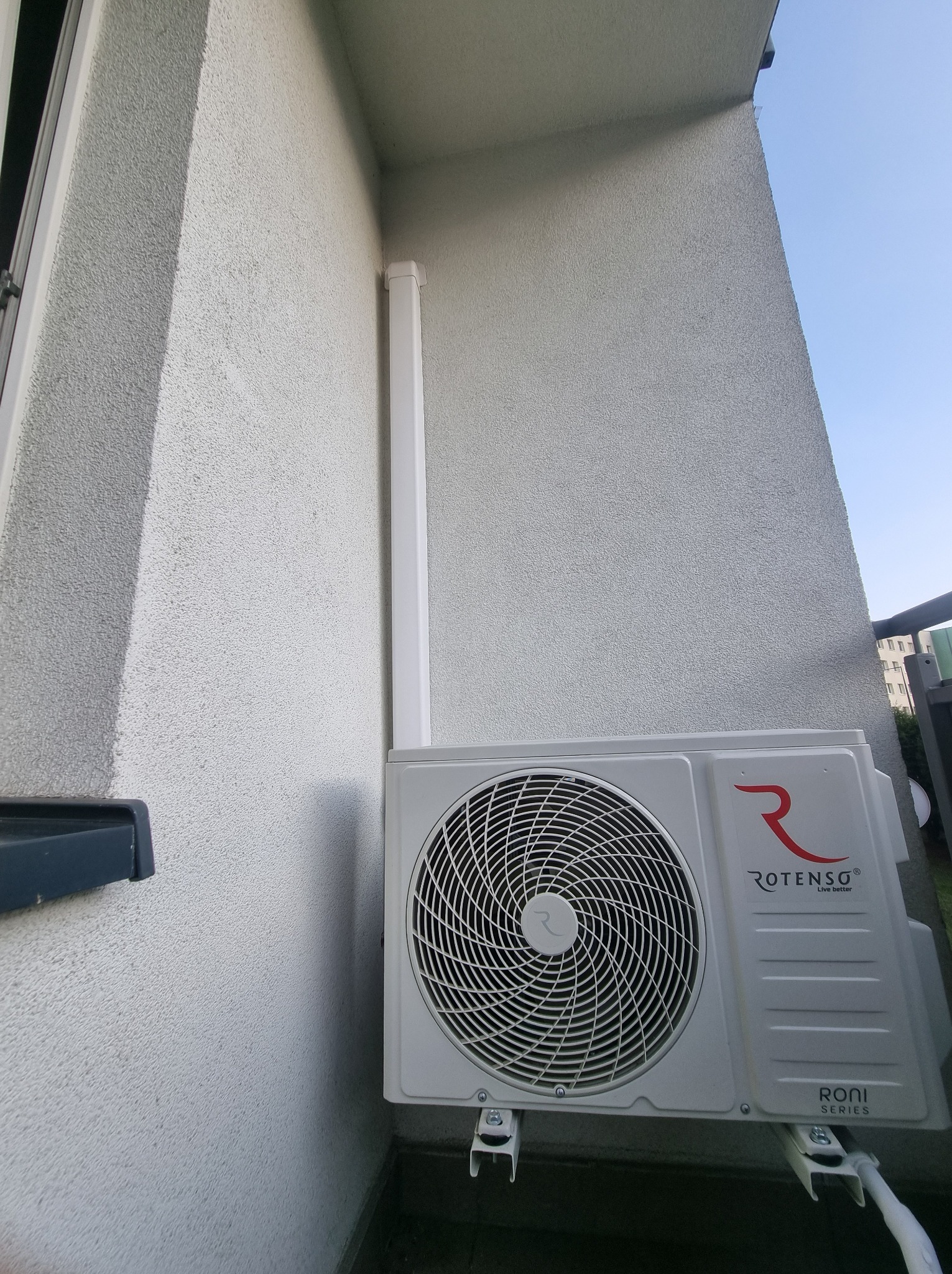 Zewnętrzna jednostka klimatyzacji Rotenso Roni Series zamontowana na elewacji budynku z widoczną instalacją odprowadzającą skropliny. Ujęcie z dołu.