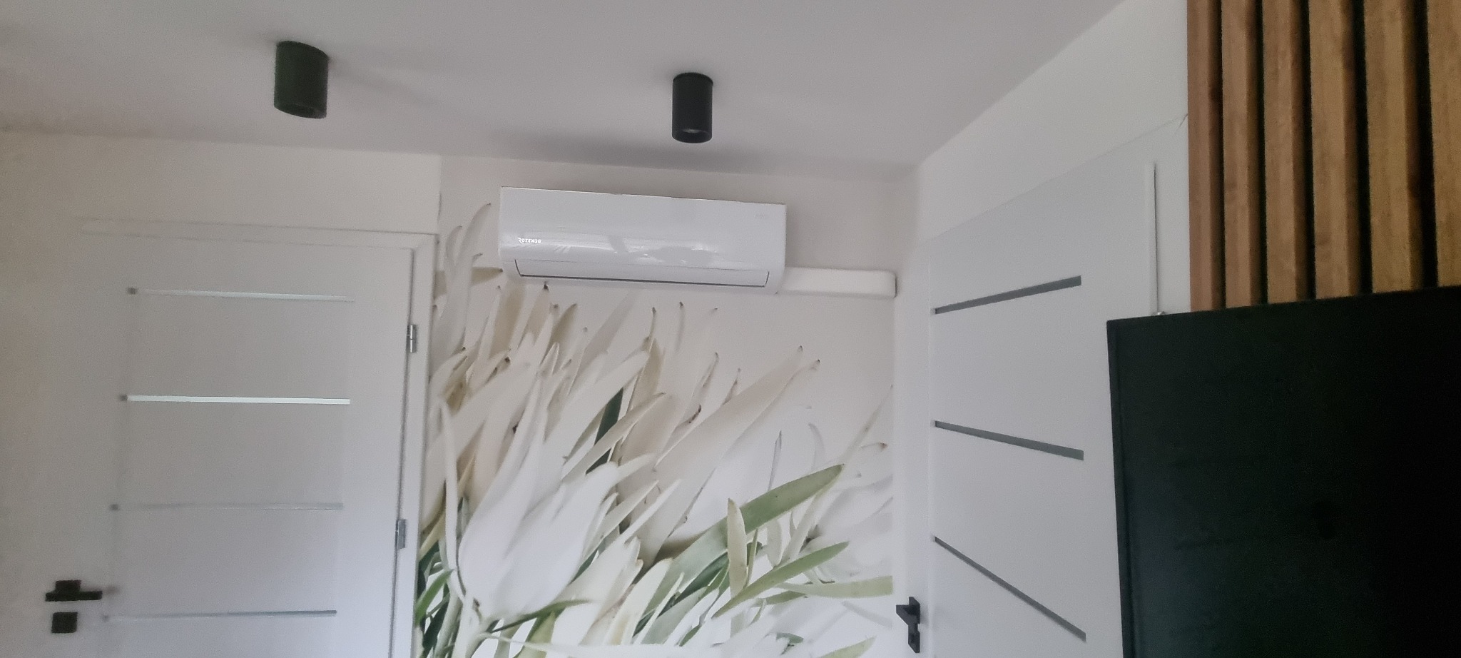Biała klimatyzacja ścienna marki Aux w nowoczesnym wnętrzu z tapetą w kwiaty i elementami dekoracyjnymi z drewna. Widoczne czarne lampy sufitowe.