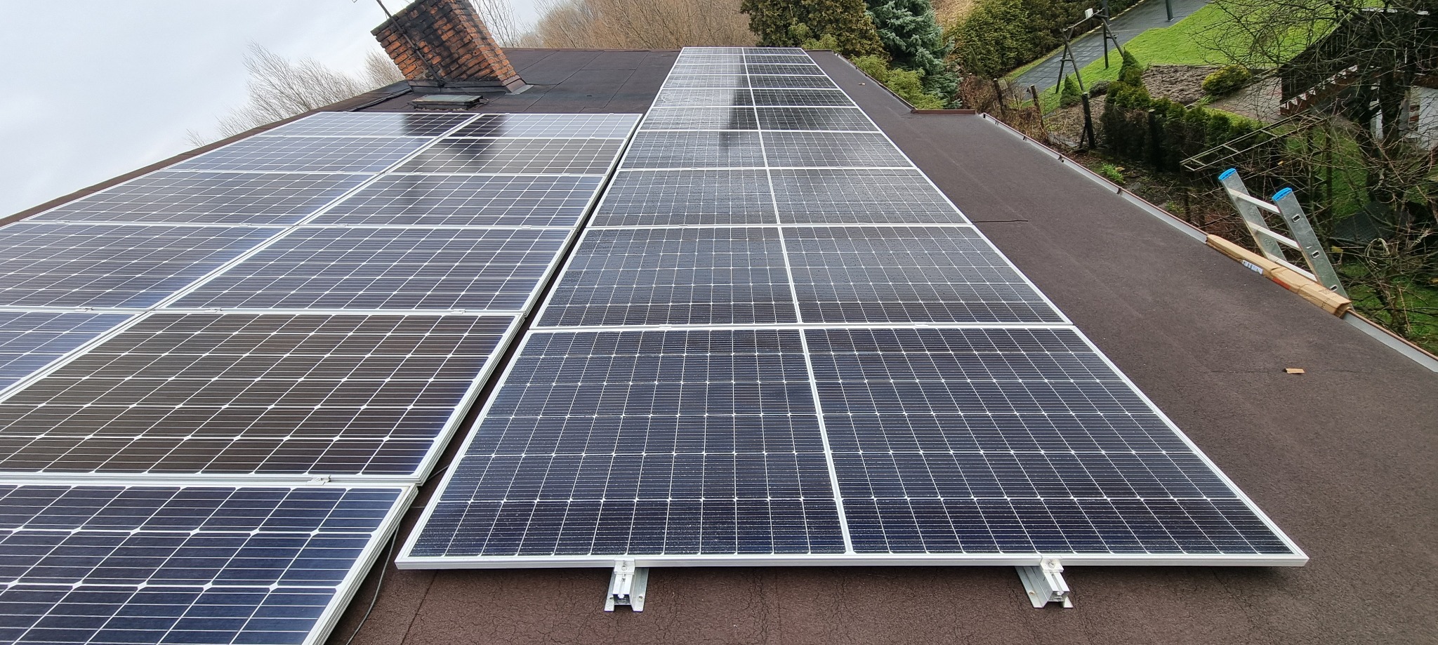 Panel fotowoltaiczny na dachu pokrytym papą, z widoczną drabiną i kominem w tle. Widok ukończonej instalacji z bliska, w pochmurny dzień.