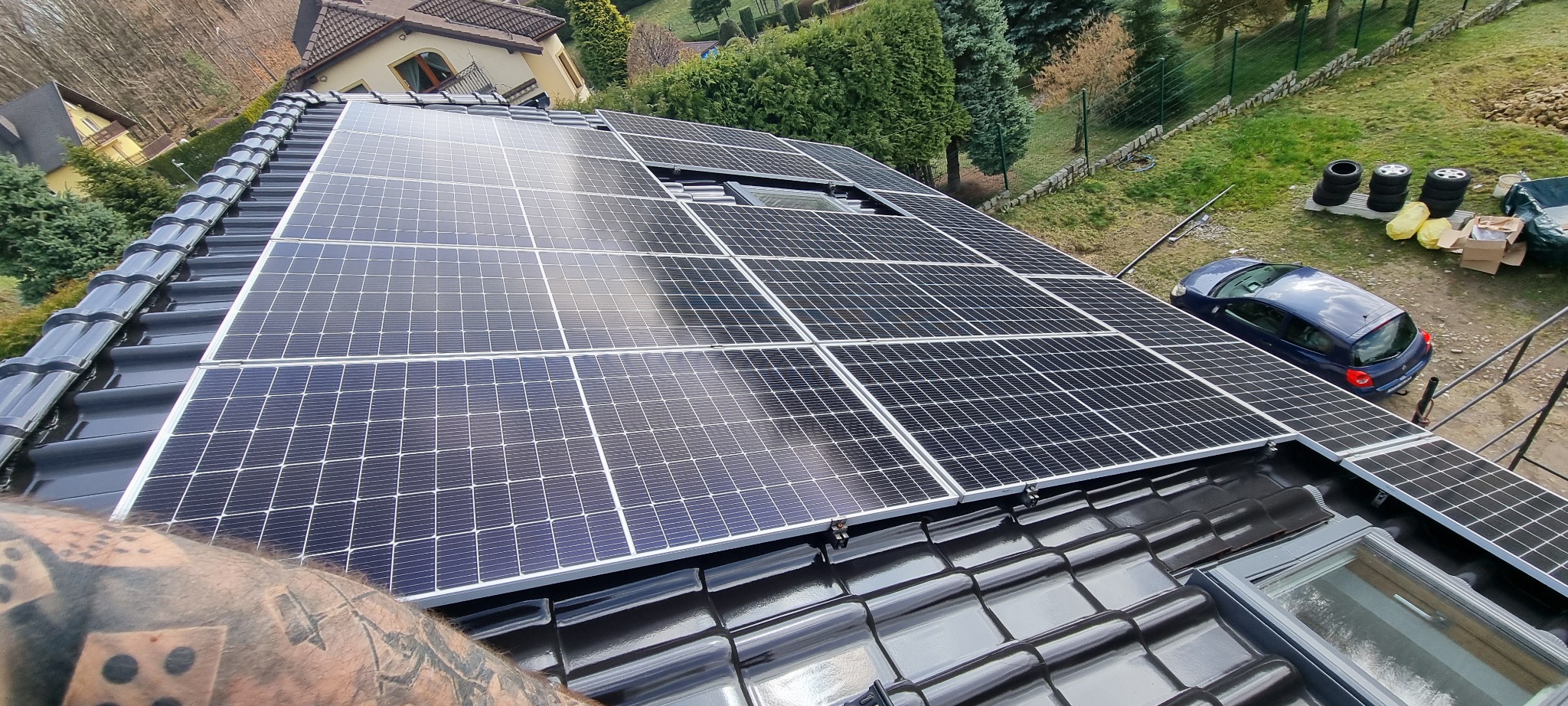 Czarny dach z panelami fotowoltaicznymi w słoneczny dzień, widok z góry, na tle domu i zieleni. W tle zaparkowany samochód i opony.