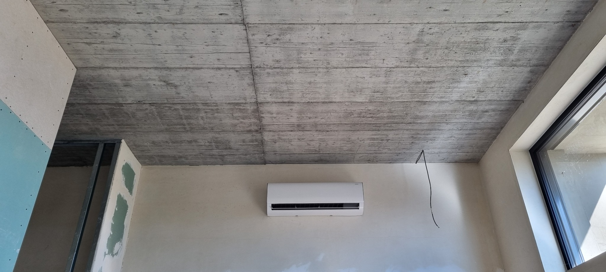 Biały klimatyzator na ścianie w surowym wnętrzu, widoczne niedokończone elementy wykończeniowe i betonowy sufit, kabel zwisający ze ściany, okno z czarną ramą.