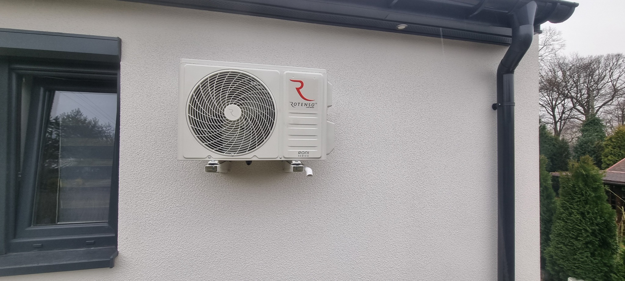 Biała jednostka zewnętrzna klimatyzacji Rotenso Roni Series zamontowana na elewacji domu z fakturą, obok okna i rynny w szarym kolorze.