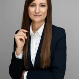 Portret kobiety w granatowej marynarce i białej koszuli na szarym tle. Uśmiechnięta, z ręką uniesioną do twarzy. Profesjonalny wizerunek doradcy finansowego.