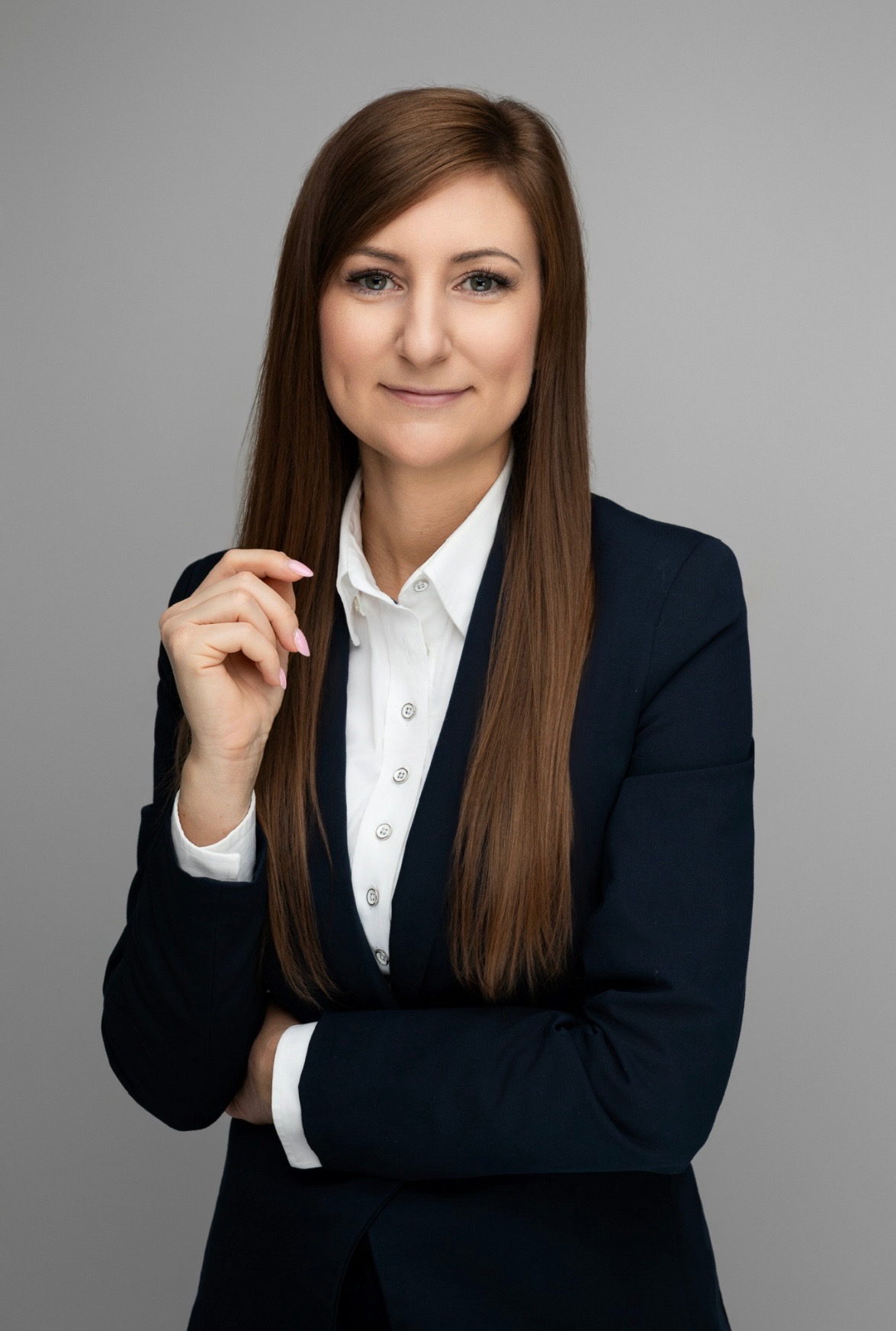 Portret kobiety w granatowej marynarce i białej koszuli na szarym tle. Uśmiechnięta, z ręką uniesioną do twarzy. Profesjonalny wizerunek doradcy finansowego.