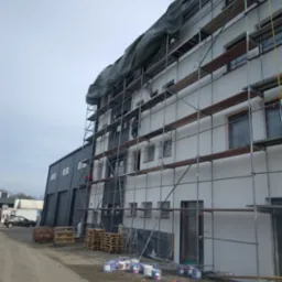 Elewacja budynku w trakcie malowania, widoczne rusztowania, farby i pracownicy. Okna zabezpieczone folią, przygotowanie do malowania.