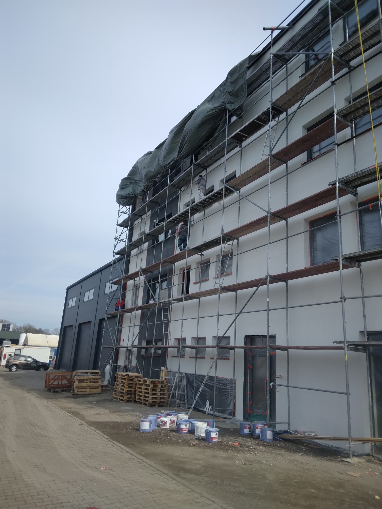 Elewacja budynku w trakcie malowania, widoczne rusztowania, farby i pracownicy. Okna zabezpieczone folią, przygotowanie do malowania.