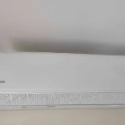 Biała klimatyzacja AUX na ścianie, widoczny panel przedni z otworami wentylacyjnymi. Minimalistyczny design, jasne wnętrze.