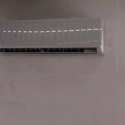 KLIMA-FLOR Łukasz Florczak - Zamontowana, biała klimatyzacja ścienna w minimalistycznym wnętrzu z szarą ścianą. Widoczne poziome żaluzje wlotu powietrza.