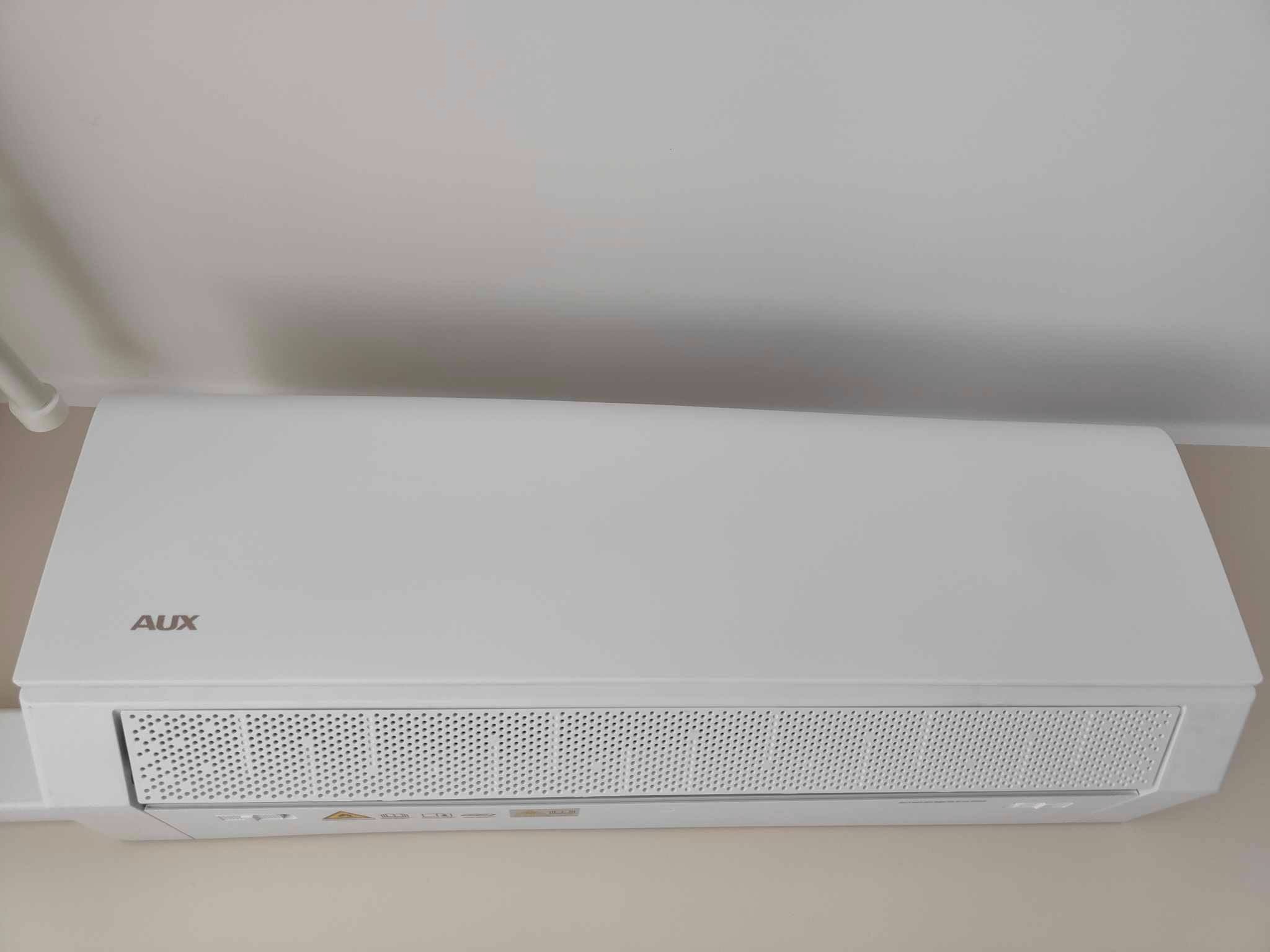 Biała klimatyzacja AUX na ścianie, widoczny panel przedni z otworami wentylacyjnymi. Minimalistyczny design, jasne wnętrze.