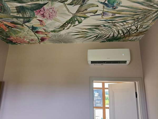 Biała klimatyzacja ścienna w pokoju z tropikalną tapetą na suficie, umieszczona nad otwartymi drzwiami. Widoczny fragment wnętrza z oknem.