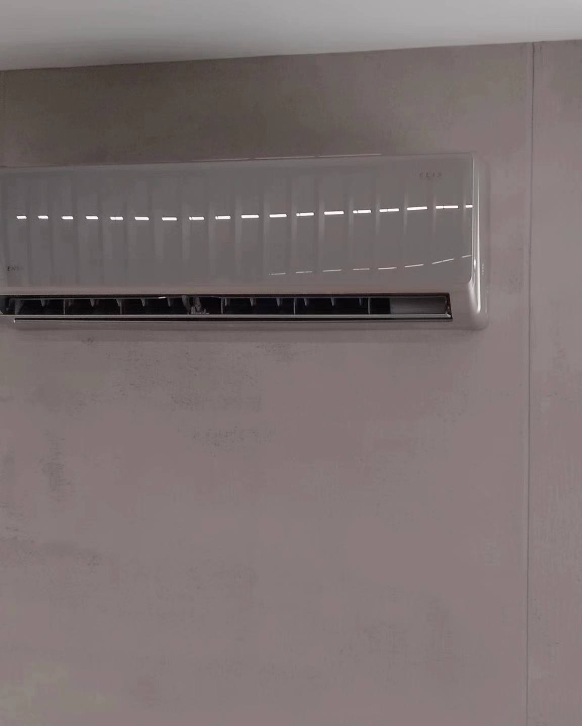 Zamontowana, biała klimatyzacja ścienna w minimalistycznym wnętrzu z szarą ścianą. Widoczne poziome żaluzje wlotu powietrza.