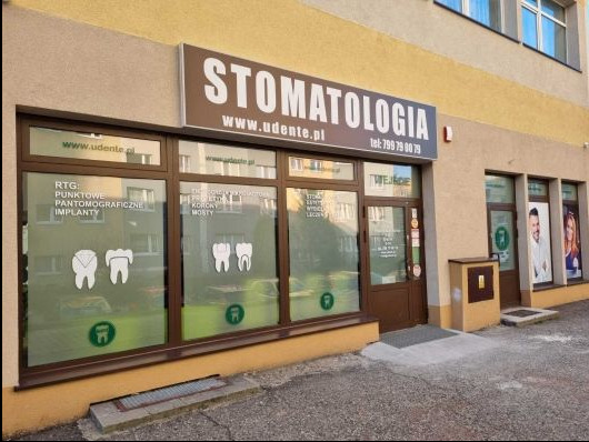 Fasada budynku stomatologii z dużym szyldem, oknami z grafikami zębów i napisem 'Wejście', oraz tabliczką z numerem telefonu.