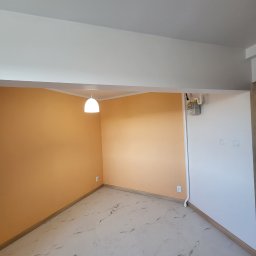 DEMIAN MISHCHUK Demli-BUD - Pomalowane na pomarańczowo ściany w pustym pokoju z marmurową podłogą i białym sufitem z lampą. Widoczny licznik gazu i gniazdka elektryczne.