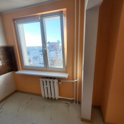 DEMIAN MISHCHUK Demli-BUD - Jasne wnętrze po remoncie: pomarańczowe ściany, białe okno z widokiem na miasto, marmurowa podłoga i grzejnik. Widoczne rury grzewcze i fragment brązowych płytek.