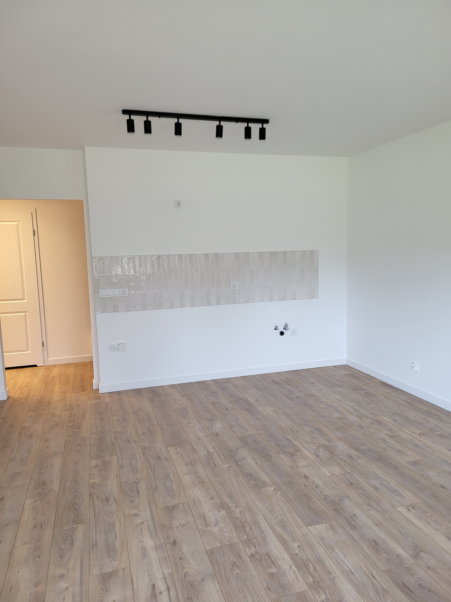 Jasne panele podłogowe i świeżo położone płytki nad blatem w pustym, odnowionym pomieszczeniu z białymi ścianami i czarną lampą sufitową.