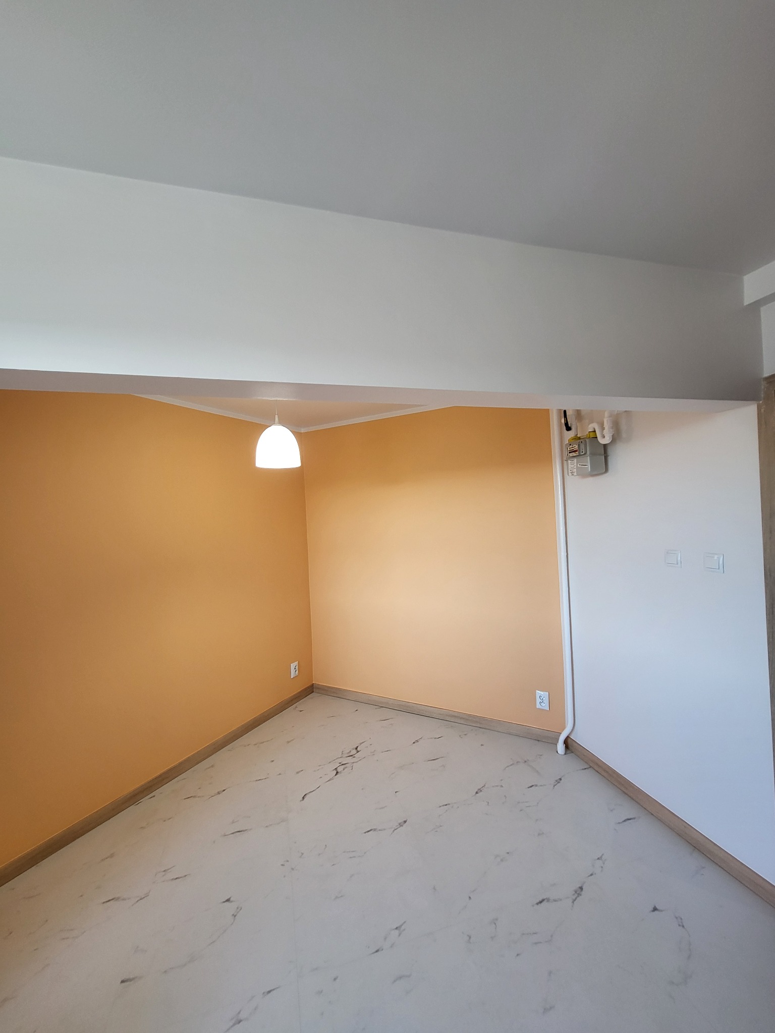 Pomalowane na pomarańczowo ściany w pustym pokoju z marmurową podłogą i białym sufitem z lampą. Widoczny licznik gazu i gniazdka elektryczne.
