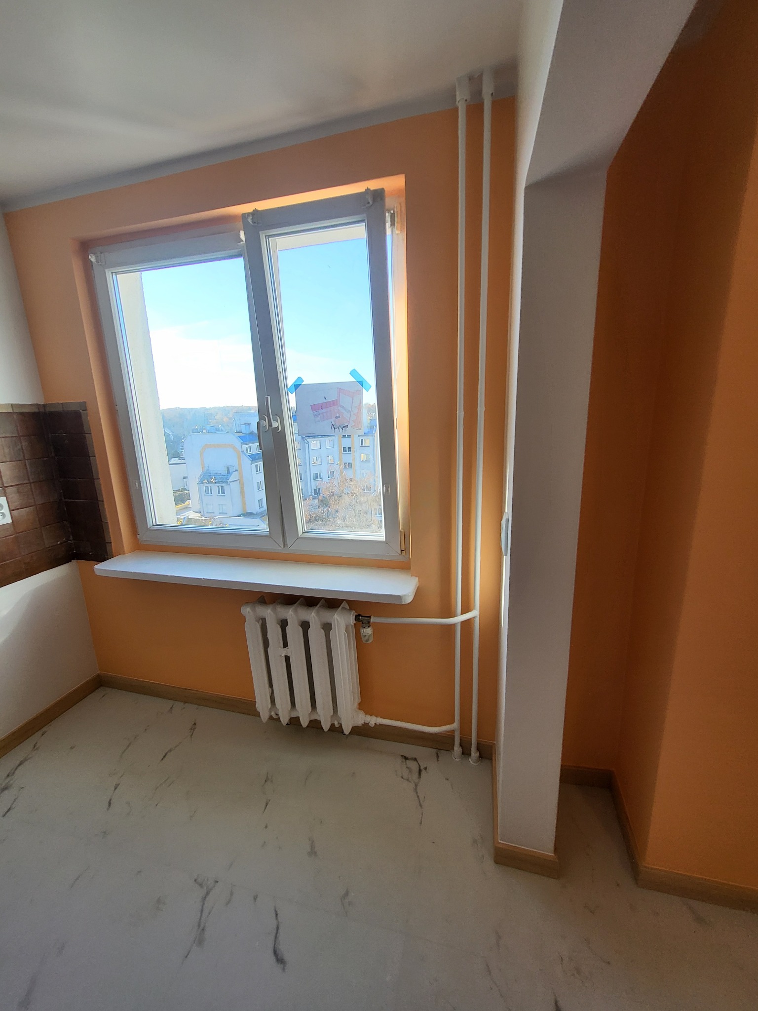 Jasne wnętrze po remoncie: pomarańczowe ściany, białe okno z widokiem na miasto, marmurowa podłoga i grzejnik. Widoczne rury grzewcze i fragment brązowych płytek.