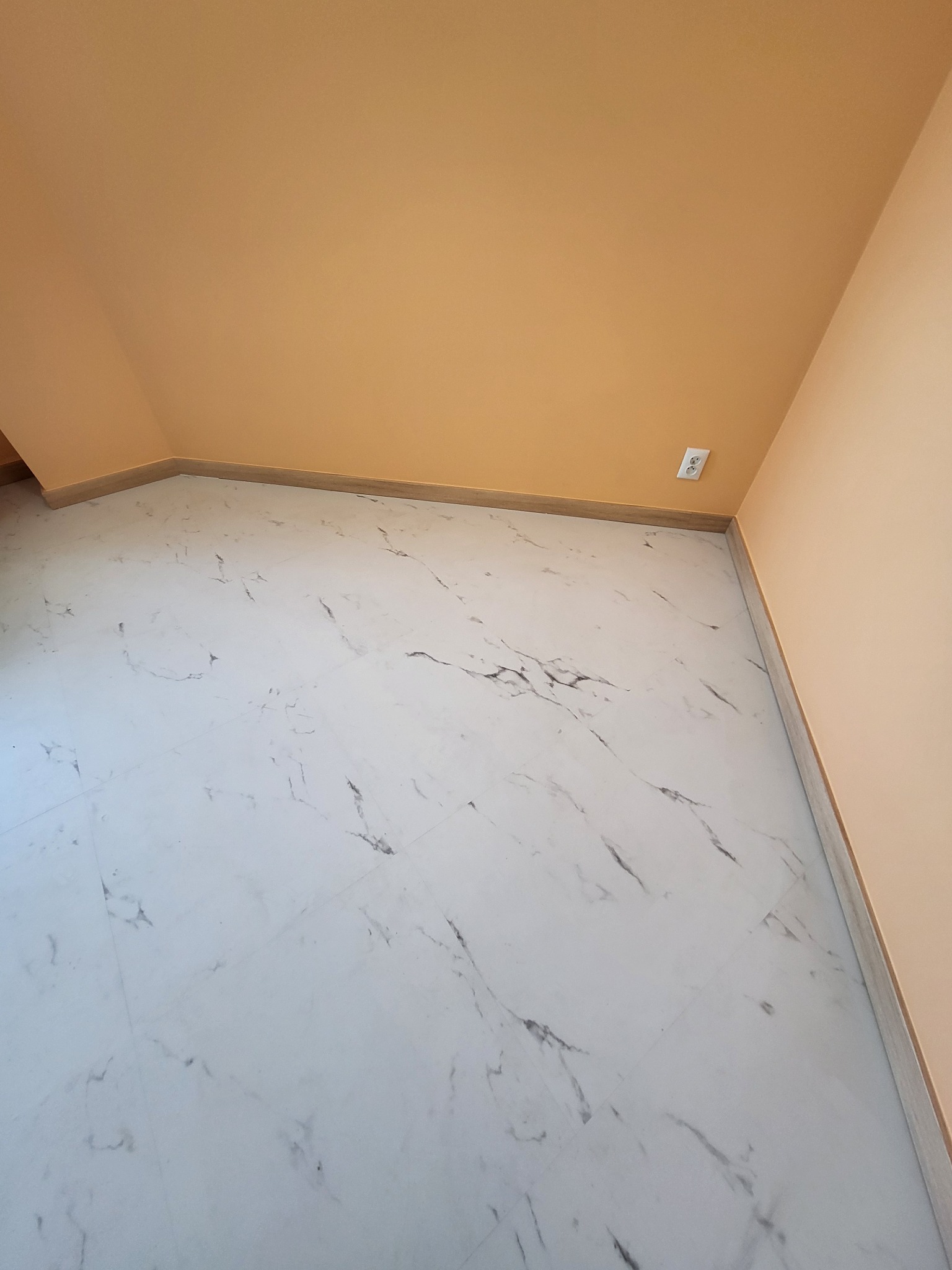Jasne wnętrze po remoncie: marmurowe płytki podłogowe z szarymi żyłkami, pomarańczowe ściany i gniazdko elektryczne w rogu. Minimalistyczny design.