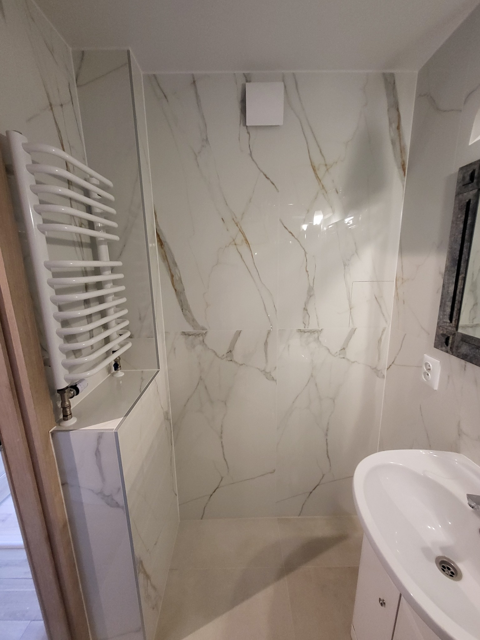 Biała łazienka z marmurowymi płytkami, grzejnikiem drabinkowym i umywalką. Widoczny fragment lustra i kratka wentylacyjna. Minimalistyczny design, jasne oświetlenie.