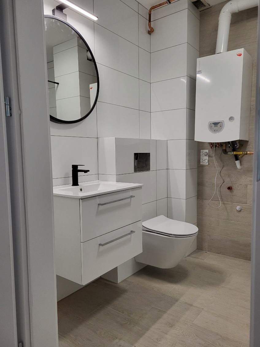 Nowoczesna łazienka z białymi płytkami, wiszącą toaletą i szafką z umywalką. Okrągłe lustro z czarną ramą i piec gazowy na ścianie. Minimalistyczny design.