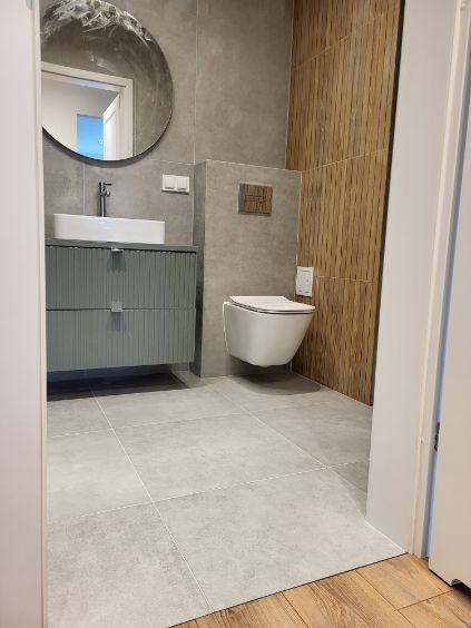 Nowoczesna łazienka z szarą armaturą, drewnianą ścianą i okrągłym lustrem. Umywalka nablatowa na szafce z frezowanymi frontami. Minimalistyczna toaleta podwieszana.