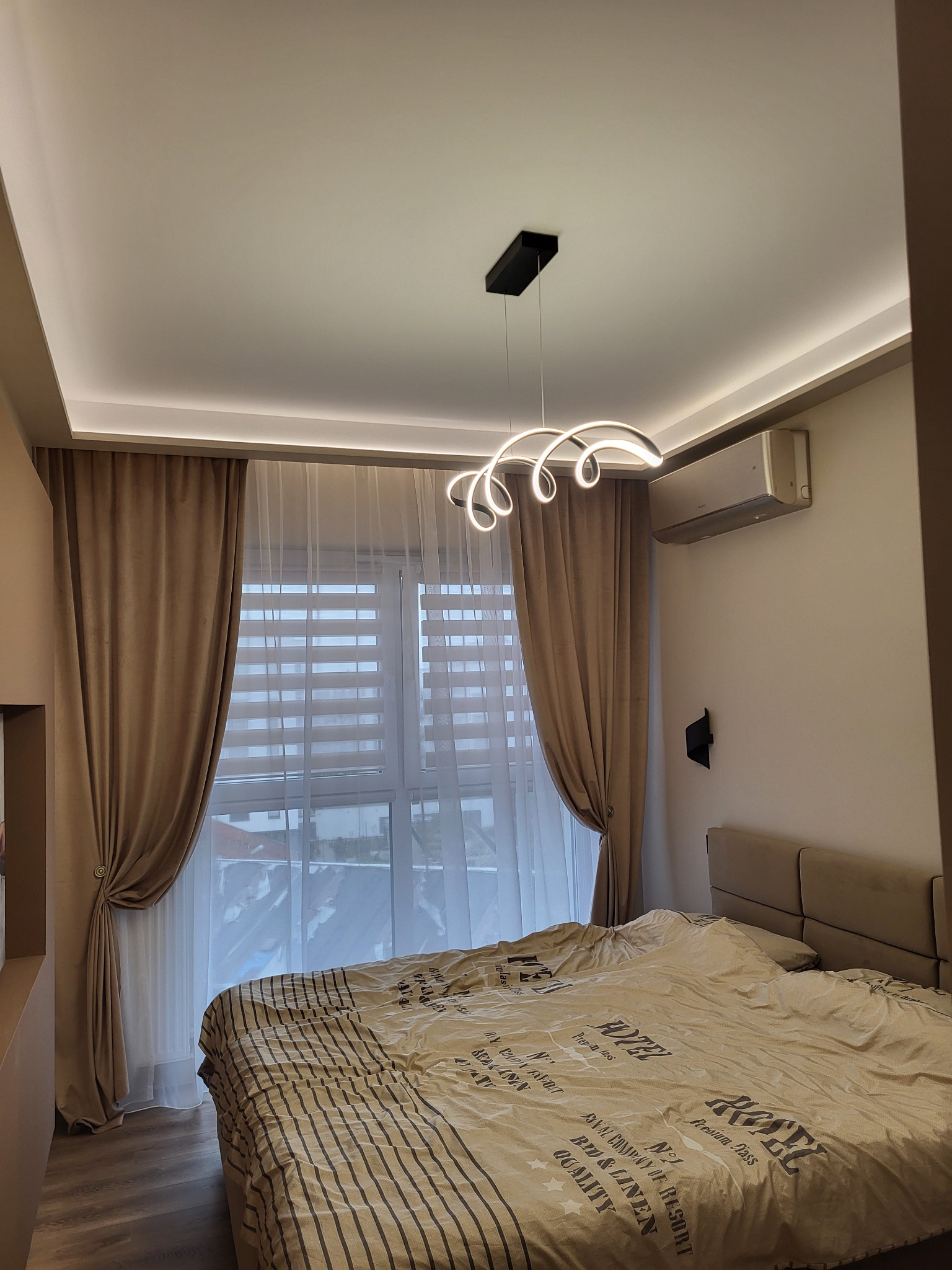 Nowoczesna sypialnia z beżowymi zasłonami, designerską lampą i klimatyzacją. Widoczne subtelne oświetlenie LED na suficie i tekstylia w stonowanych barwach.