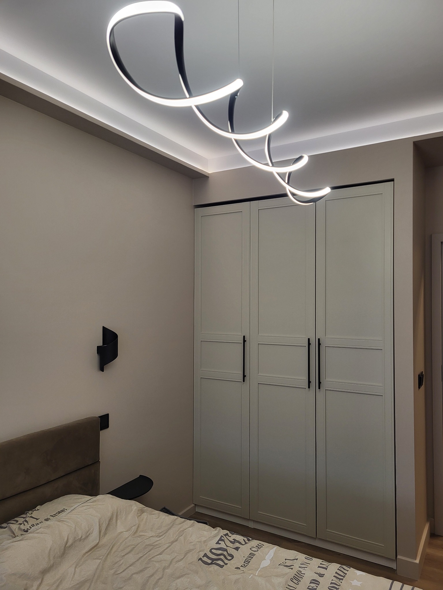 Sypialnia po remoncie: biała szafa w zabudowie, designerska lampa LED, listwy LED pod sufitem, beżowe ściany, łóżko z brązowym zagłówkiem i czarnymi lampkami.