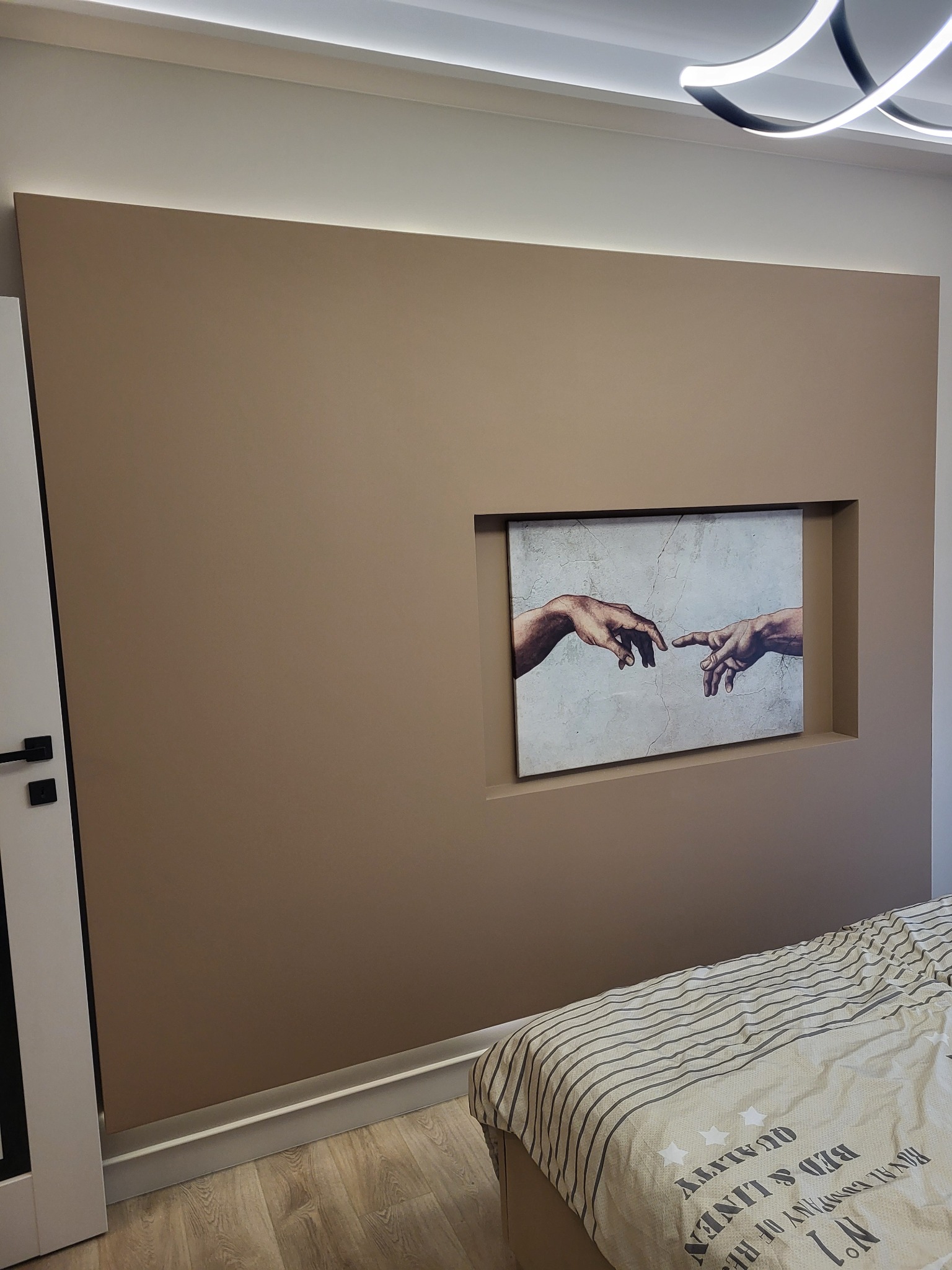 Beżowa ściana z wnęką, w której umieszczono obraz 'Stworzenie Adama'. Nowoczesne oświetlenie sufitowe i fragment łóżka w dolnym rogu kadru.