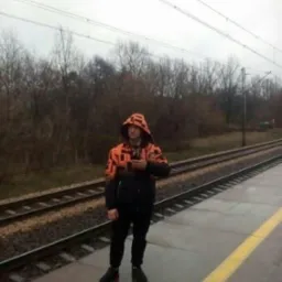 Osoba w pomarańczowej kurtce z kapturem stoi na peronie kolejowym, trzymając telefon. W tle widoczne tory kolejowe i drzewa bez liści w pochmurny dzień.