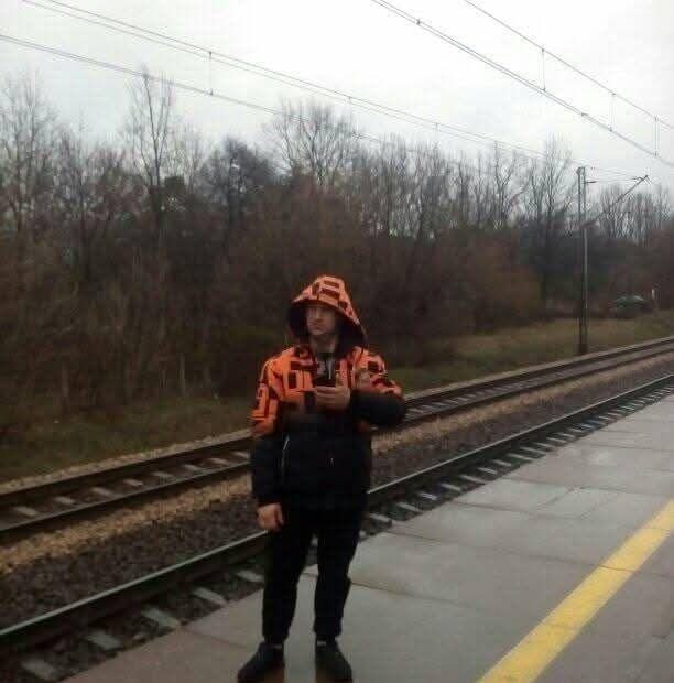 Osoba w pomarańczowej kurtce z kapturem stoi na peronie kolejowym, trzymając telefon. W tle widoczne tory kolejowe i drzewa bez liści w pochmurny dzień.