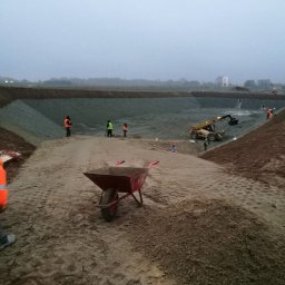 Kamil Świątek SK-Tech - Niwelacja terenu pod budowę zbiornika retencyjnego. W tle pracujący sprzęt budowlany i ekipa w odblaskowych kamizelkach. Na pierwszym planie taczka.