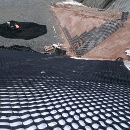 Kamil Świątek SK-Tech - Układanie geokraty na podłożu gruntowym. Widoczne elementy mocujące i pracownicy w odblaskowych kamizelkach. Etap przygotowania terenu pod budowę.