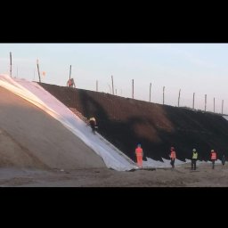 Kamil Świątek SK-Tech - Prace ziemne: robotnicy w odblaskowych ubraniach układają białą geowłókninę na stoku wzmocnionym siatką. W tle widoczne drewniane pale.