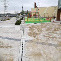 Układanie kostki brukowej na placu zabaw z widocznym systemem drenażowym i elementami placu zabaw w tle. Prace wykończeniowe, piasek na kostce.