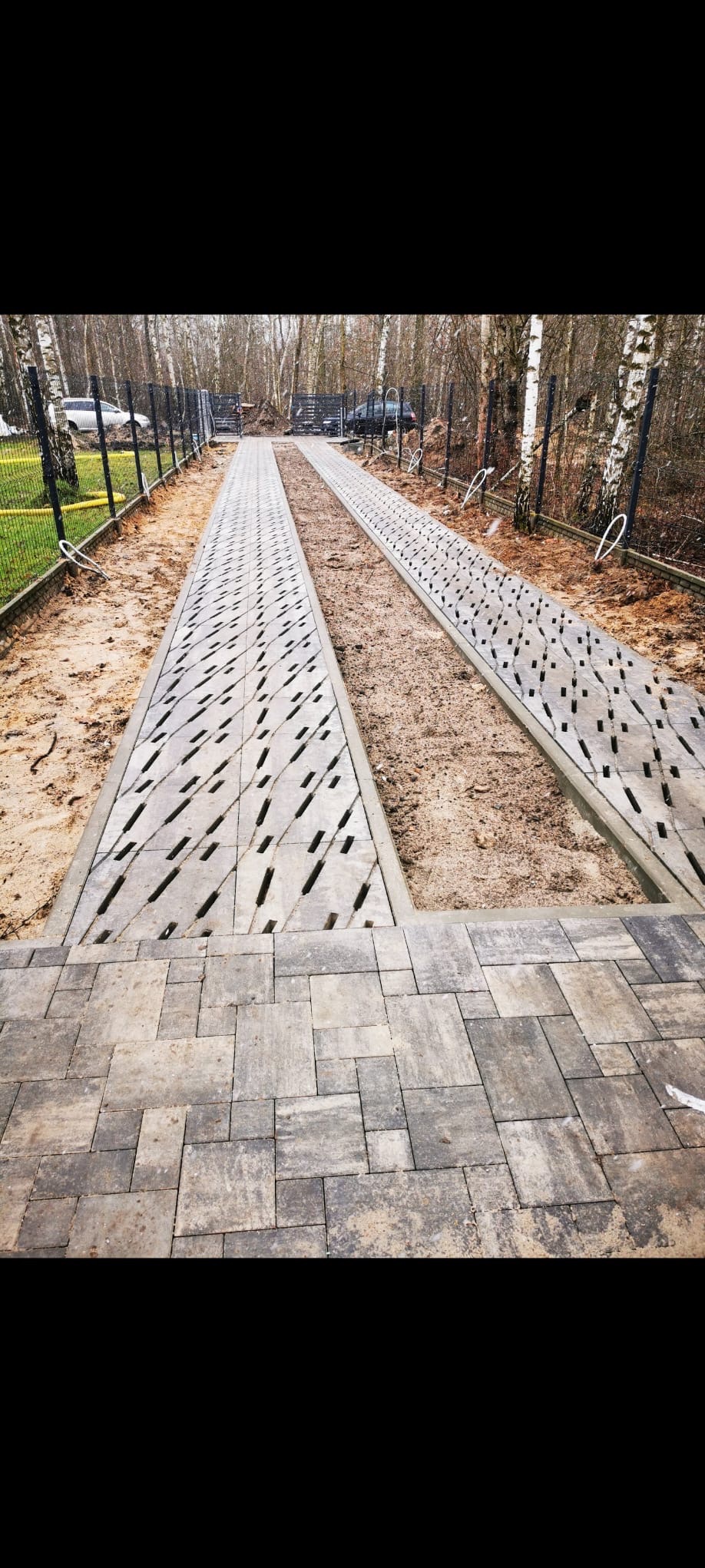 Wykończona ścieżka z kostki brukowej, z elementami drenażu liniowego, otoczona piaskiem i ogrodzeniem panelowym na tle drzew. Widok z góry, perspektywa zbieżna.