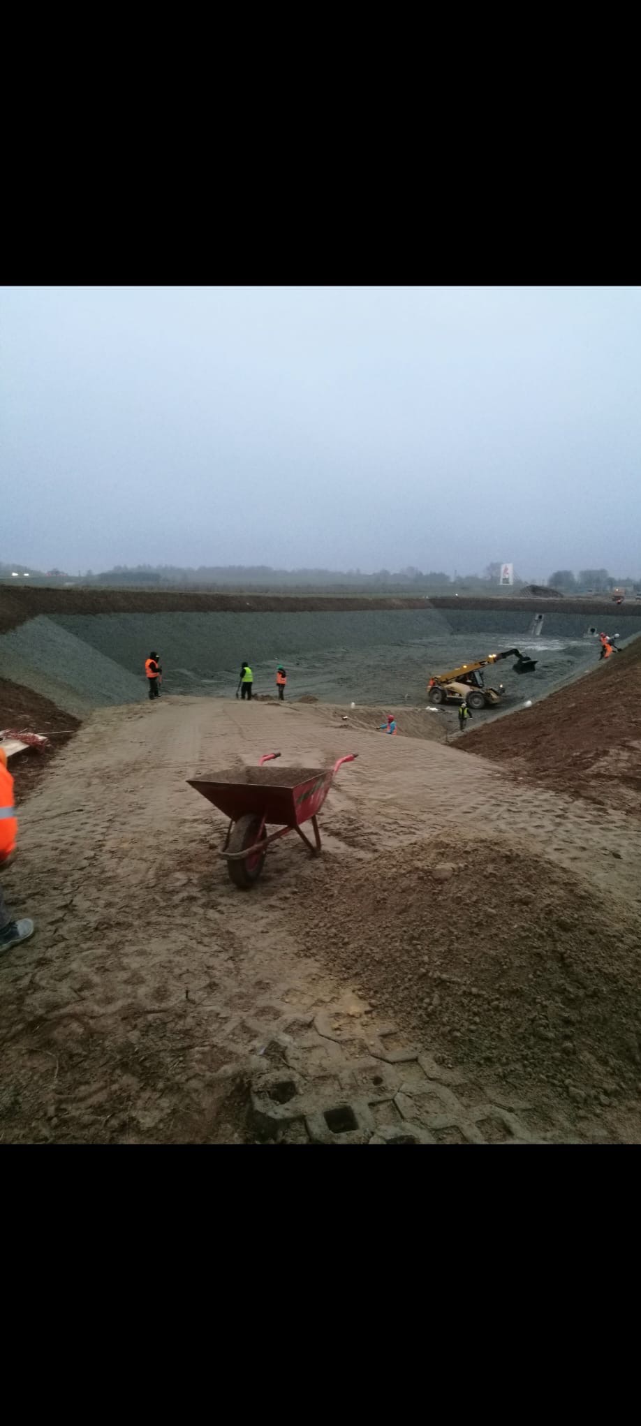 Niwelacja terenu pod budowę zbiornika retencyjnego. W tle pracujący sprzęt budowlany i ekipa w odblaskowych kamizelkach. Na pierwszym planie taczka.