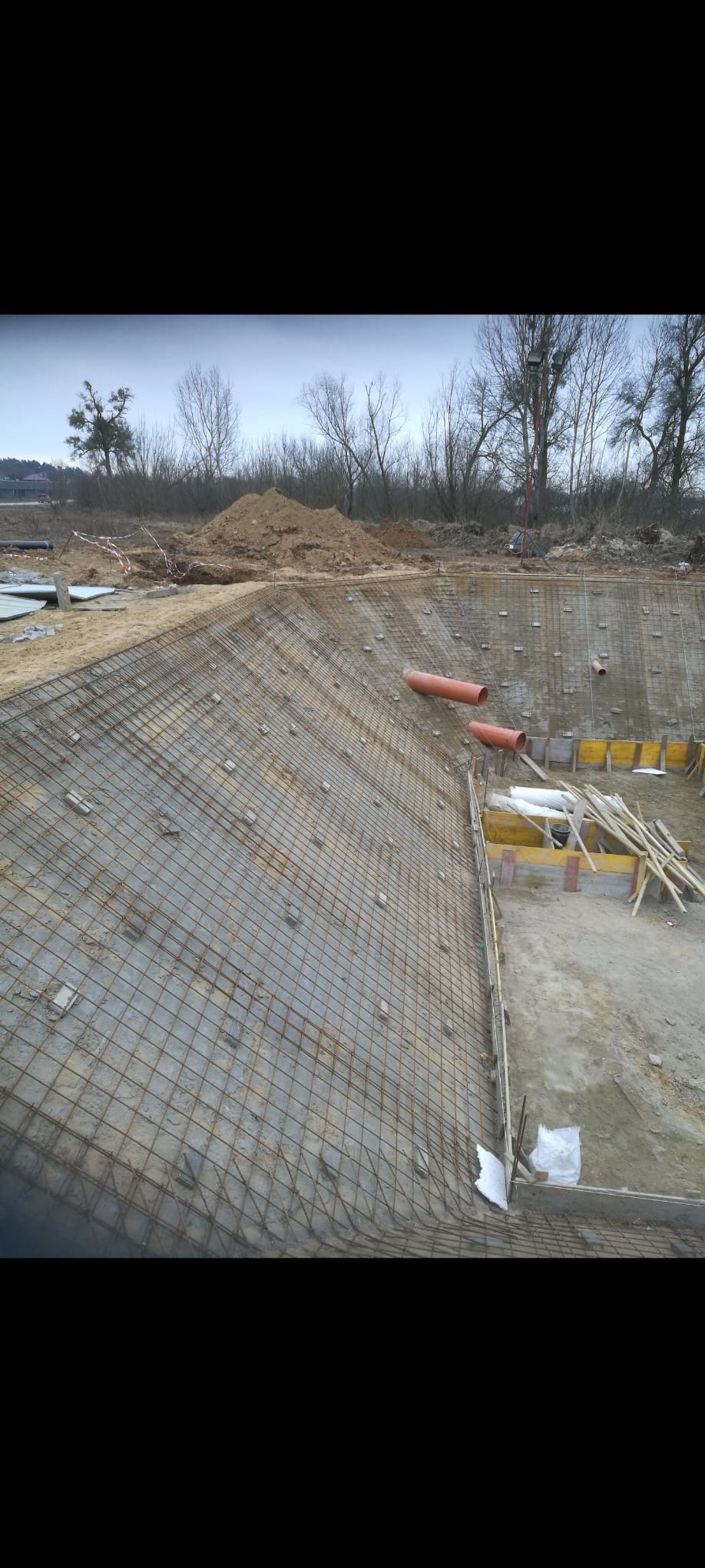 Siatka zbrojeniowa na zboczu wykopu, widoczne szalunki i rury drenażowe, w tle hałda ziemi. Prace przygotowawcze pod budowę zbiornika.