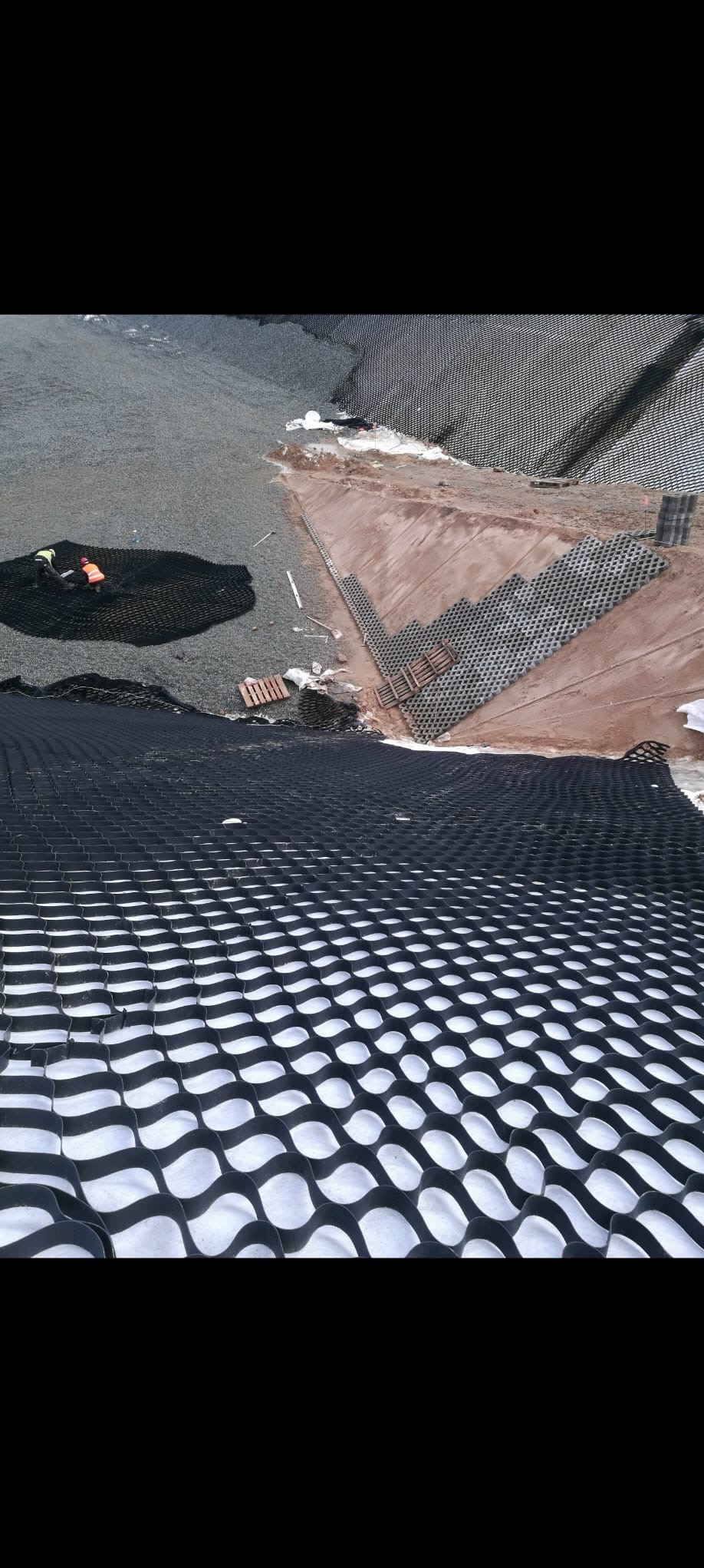 Układanie geokraty na podłożu gruntowym. Widoczne elementy mocujące i pracownicy w odblaskowych kamizelkach. Etap przygotowania terenu pod budowę.