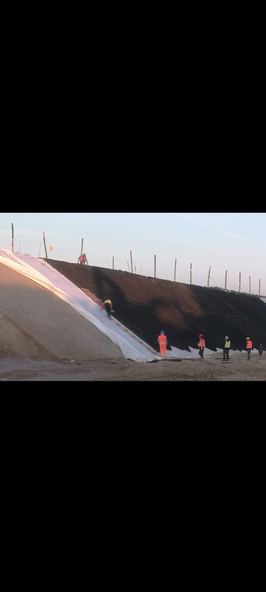 Prace ziemne: robotnicy w odblaskowych ubraniach układają białą geowłókninę na stoku wzmocnionym siatką. W tle widoczne drewniane pale.