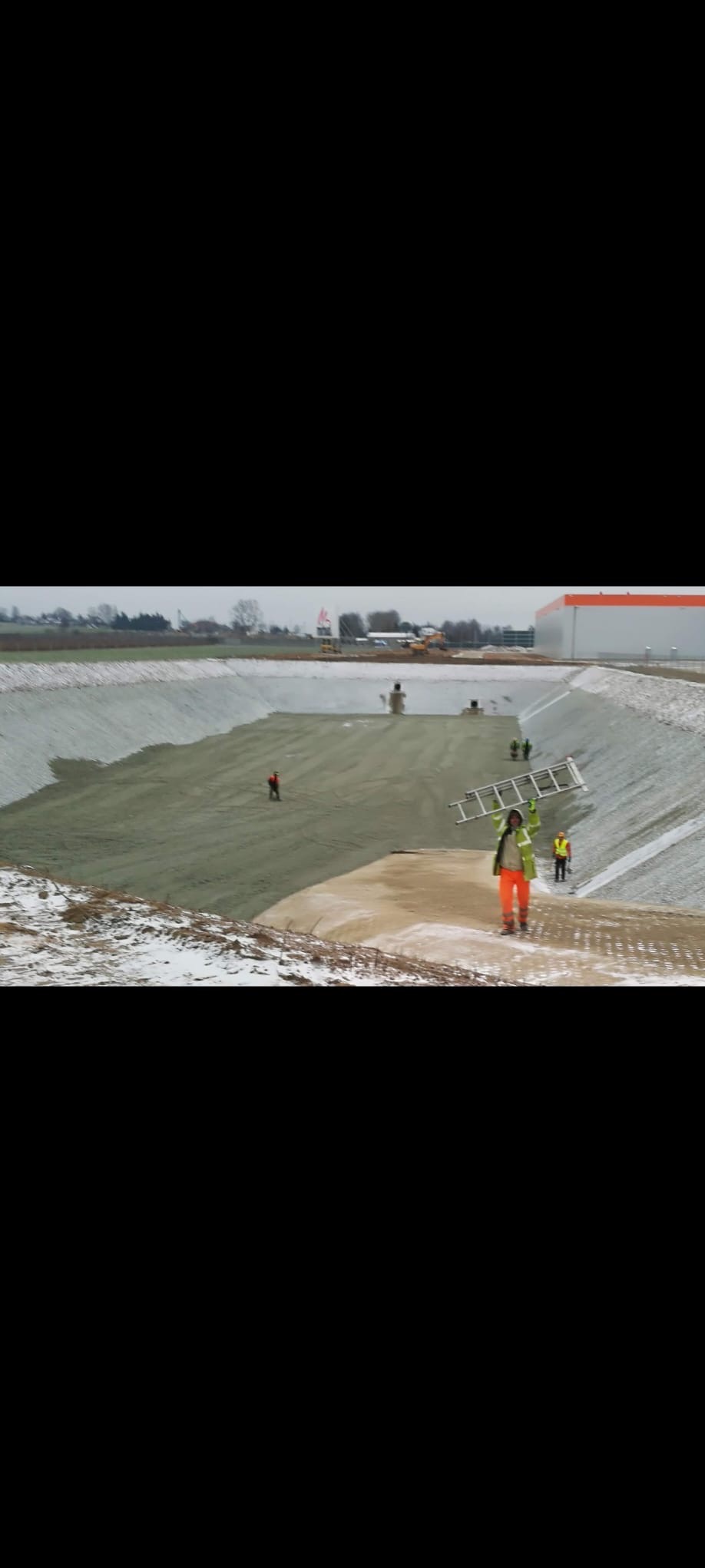 Budowa zbiornika retencyjnego: robotnicy w odblaskowych ubraniach pracują na zboczu wyłożonym zieloną geowłókniną. W tle budynek i sprzęt budowlany.