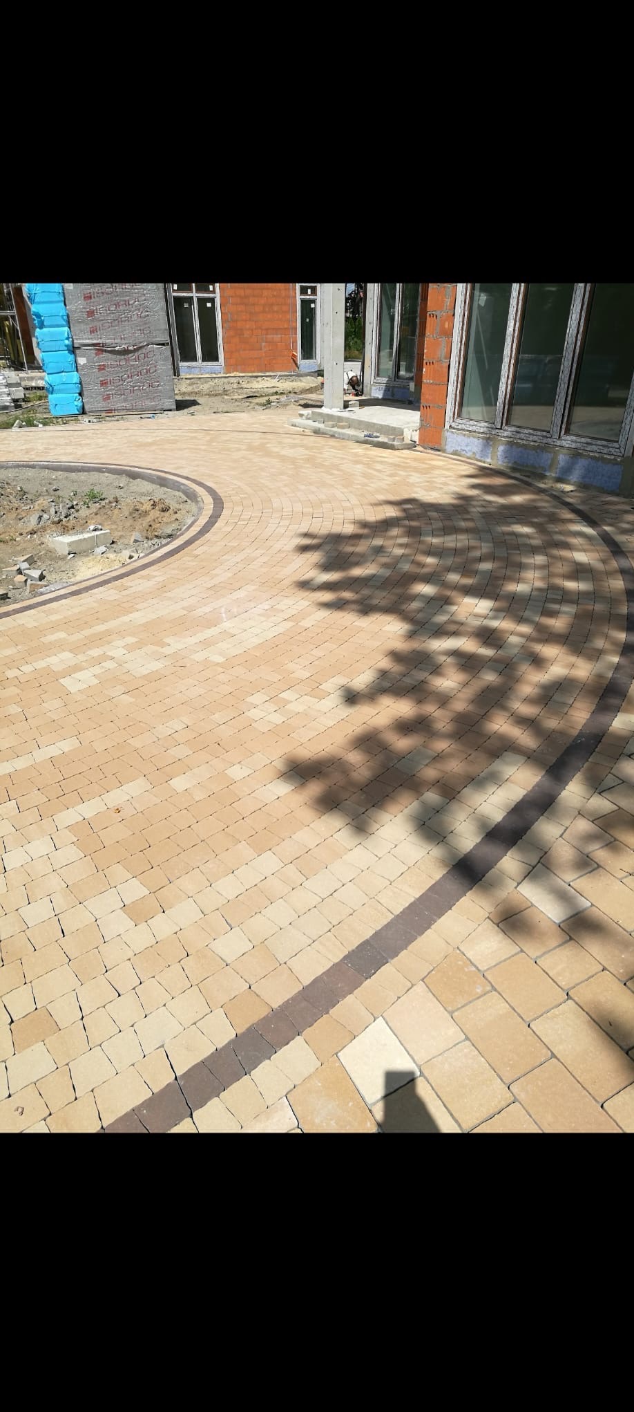 Ułożona, beżowa kostka brukowa tworząca okrągły wzór na podjeździe. Ciemniejsze obramowanie podkreśla kształt. W tle budynek w trakcie budowy.