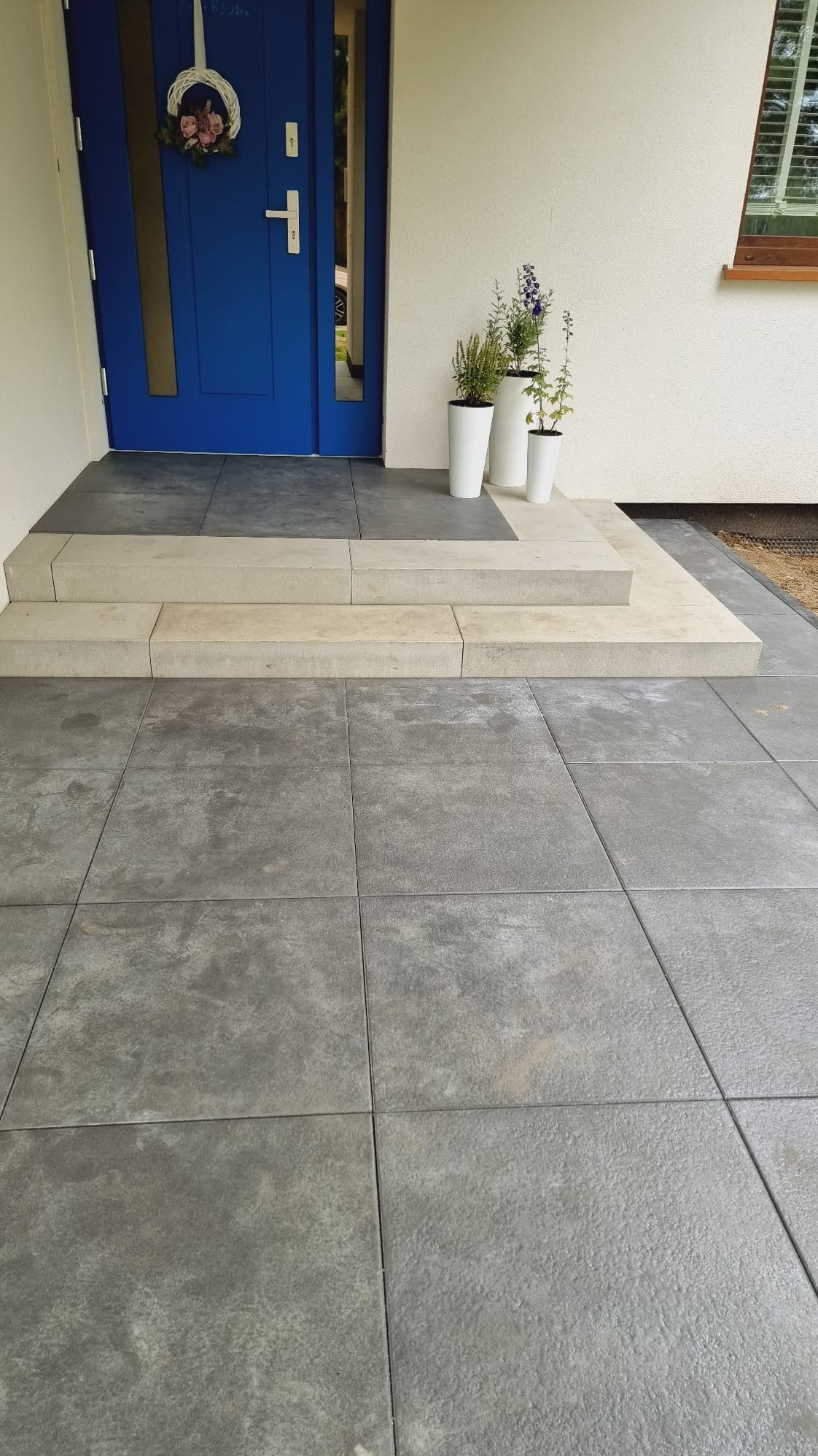 Nowoczesny taras z szarych, kwadratowych płyt, prowadzący do niebieskich drzwi wejściowych. Trzy białe donice z roślinami zdobią wejście.